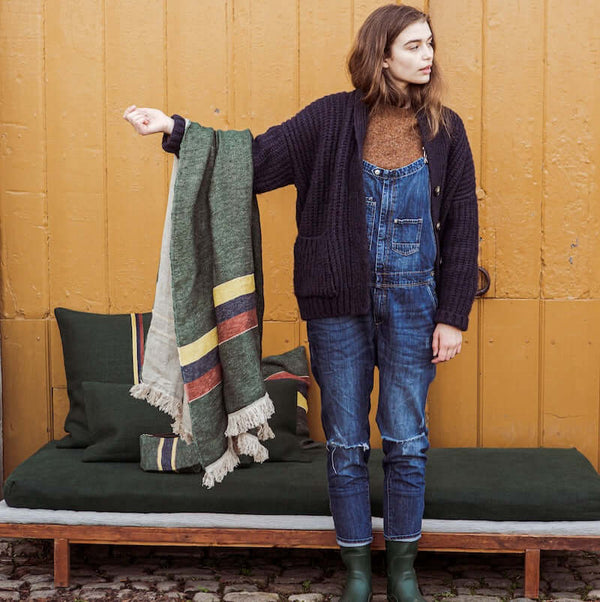 A girl holding up a gorgeous Libeco Belgian linen throw Spruce that has a green ground with stripes in gold, navy, and mahogany with flax-colored fringe.